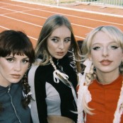 Three members of the band Blusher stood on an athletics track all with plaits and bows in their hair